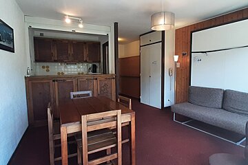 Cozy studio apartment featuring a wooden dining table, a small kitchen area, a grey sofa, and red carpeting.