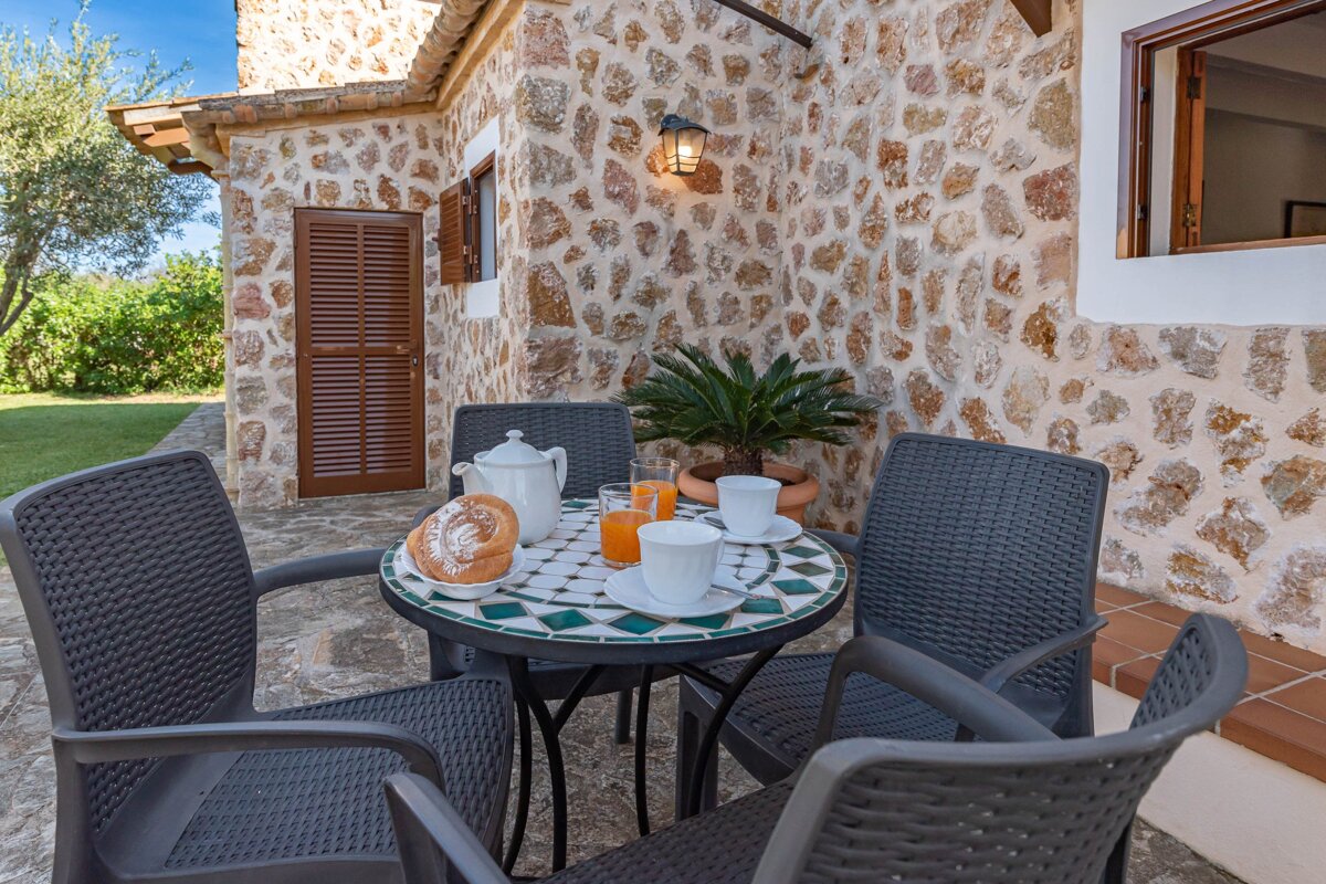 A rustic stone patio with a mosaic table set for breakfast (pastries, coffee, juice) and four chairs, against a charming stone house and green garden.