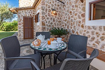 A rustic stone patio with a mosaic table set for breakfast (pastries, coffee, juice) and four chairs, against a charming stone house and green garden.