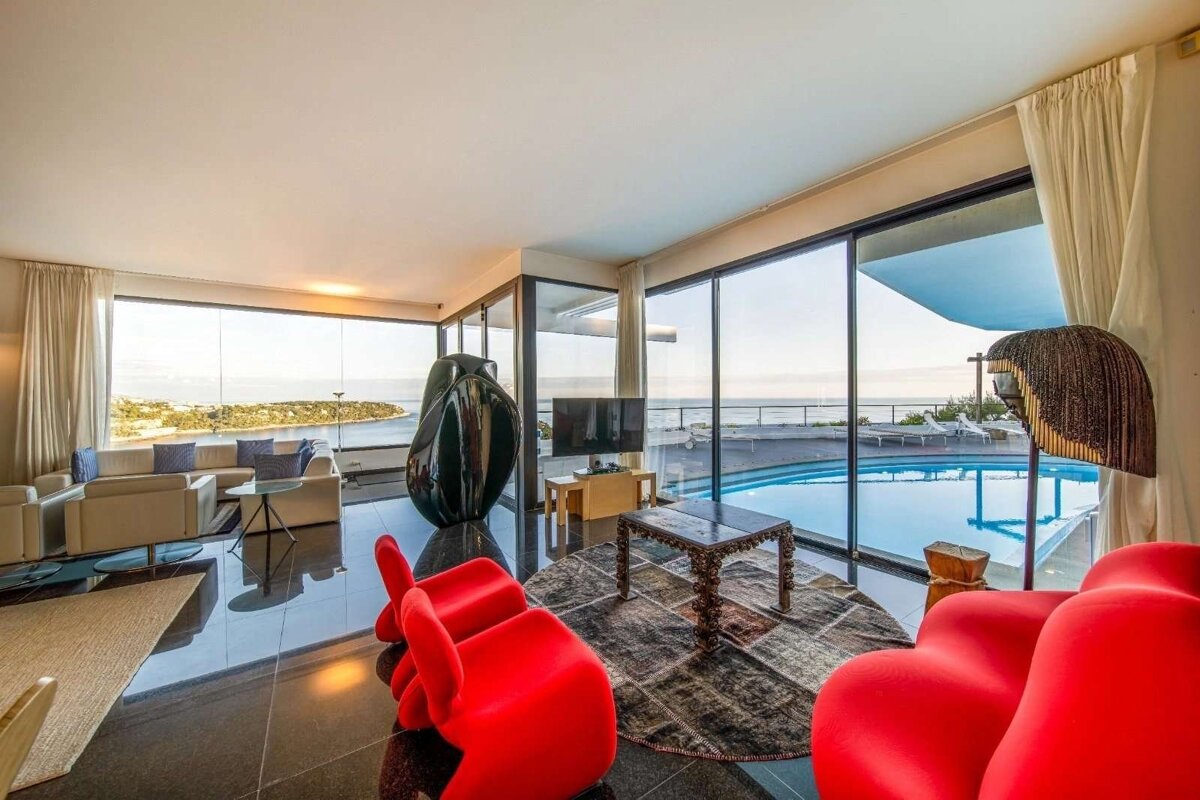 Sleek living room with vast ocean views, an infinity pool, and eclectic decor including bold red chairs and a dark abstract sculpture.