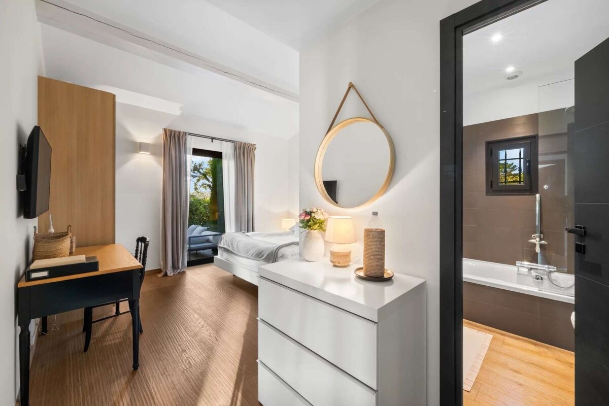 Bright bedroom with a modern bed, desk, and a dresser, featuring a glass door to a patio and an adjoining bathroom with a tub.