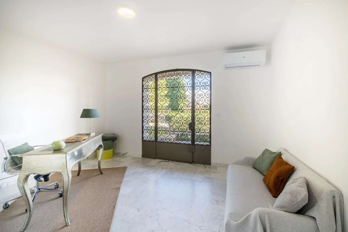 A bright, minimalist room with a light desk, sofa, and ornate wrought iron doors revealing greenery. White walls, marble floor, and AC unit.