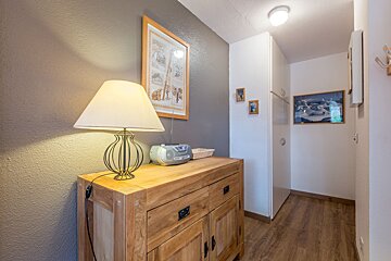 A rustic wooden cabinet with a lamp, radio, and framed art against a gray wall, leading into a brightly lit hallway with white walls and wooden flooring.
