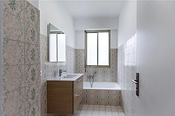 A narrow bathroom featuring patterned wall tiles, a wooden vanity with a sink and mirror, and a bathtub positioned under a window.