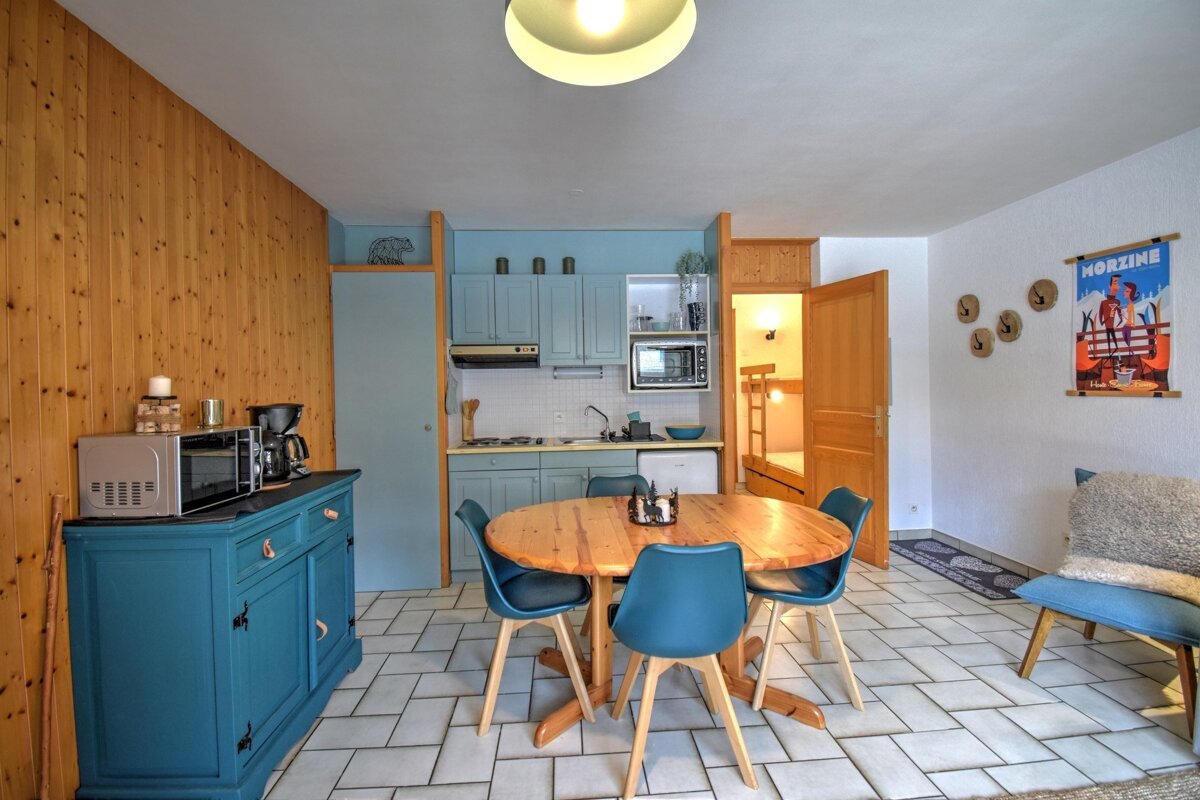 A cozy kitchen-dining room with light blue cabinets, wooden table & blue chairs, wood paneling, and tiled floor. Ski poster and bunk beds visible.