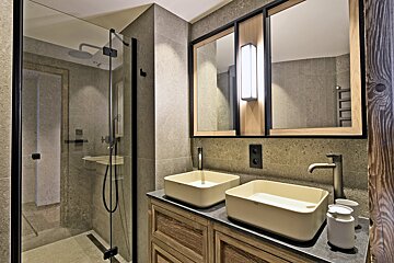 Modern bathroom with gray tiled walls, a glass shower, double cream vessel sinks on a wooden vanity, and a framed mirror with lighting.