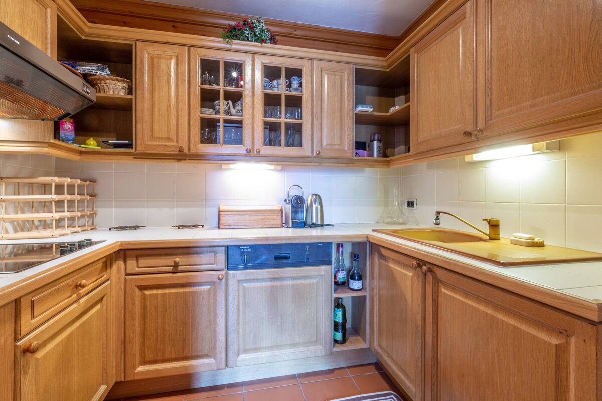 A cozy kitchen featuring light wooden cabinets, white tiled backsplash, built-in appliances, a stovetop, and a golden sink. Warm lighting creates a homey atmosphere.