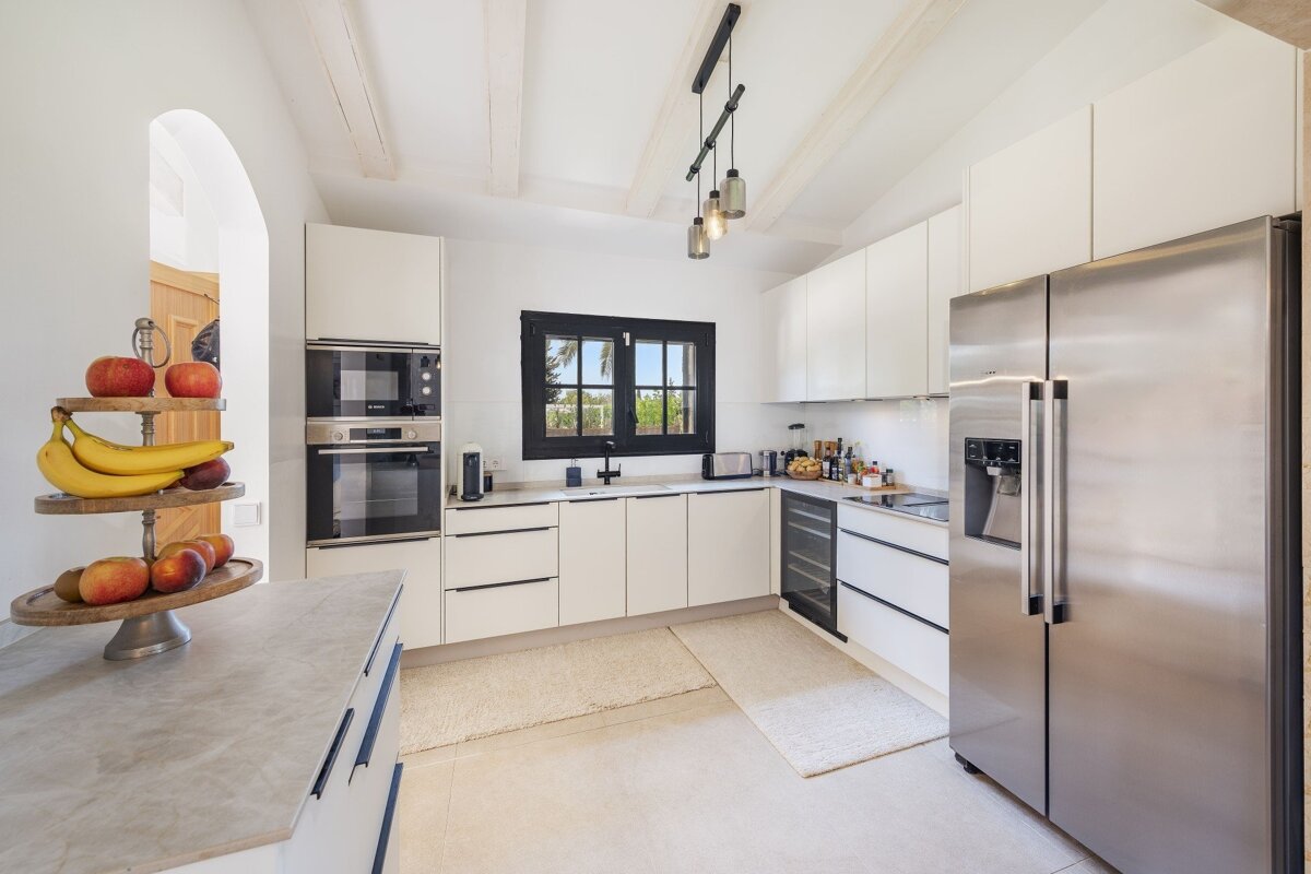 A sleek, modern white kitchen with stainless steel appliances, a black-framed window, exposed wooden beams, and a tiered fruit stand on the counter.