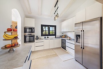 A modern, bright white kitchen features stainless steel appliances, sleek cabinets, a black-framed window, and a fruit stand. Exposed beams add charm.