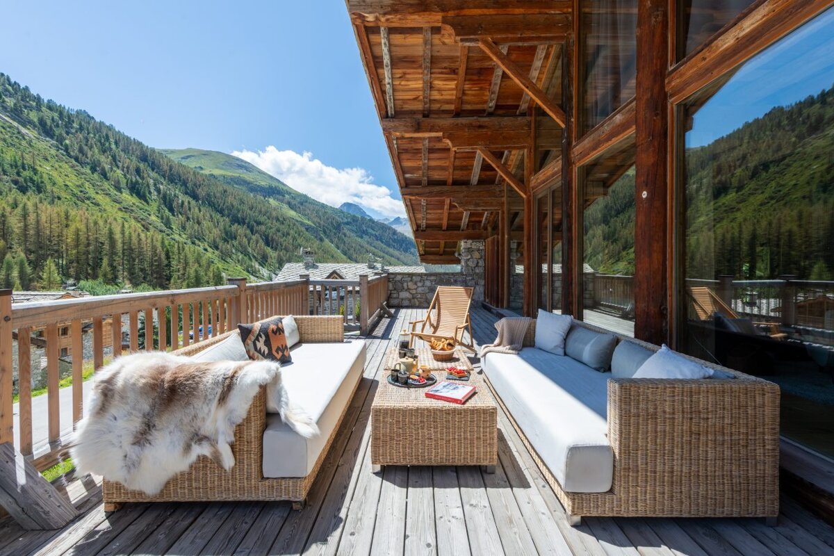 A couch with a fur blanket on it sits on a wooden deck with mountains in the background