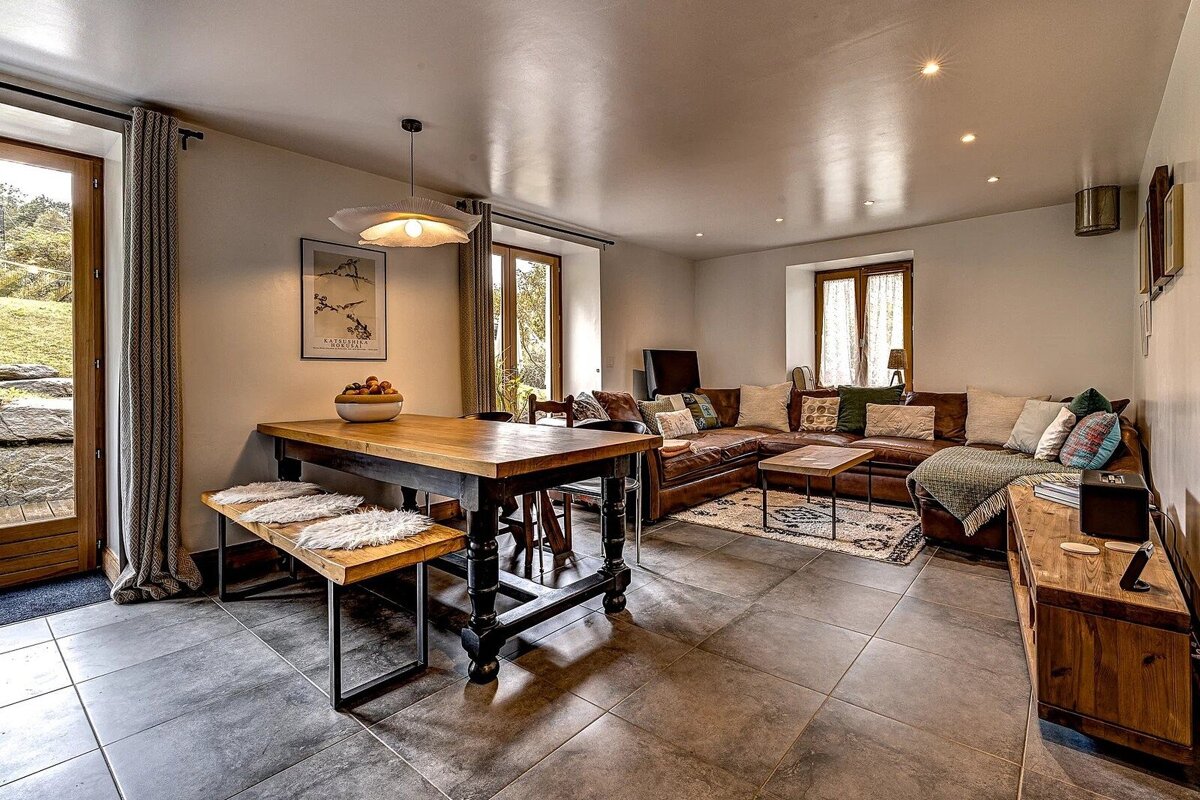 Cozy rustic-modern living space featuring a wooden dining table, tiled flooring, and a large brown leather sectional sofa.