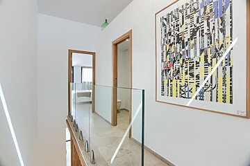 A modern, sunlit hallway features white walls, glass railings, wooden doorframes, and a large abstract artwork. Doors lead to a bedroom and bathroom.