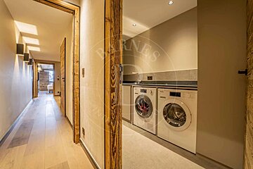A cozy hallway with wooden door frames and light floors leads to a modern laundry room with two white washing machines, a sink, and countertops.