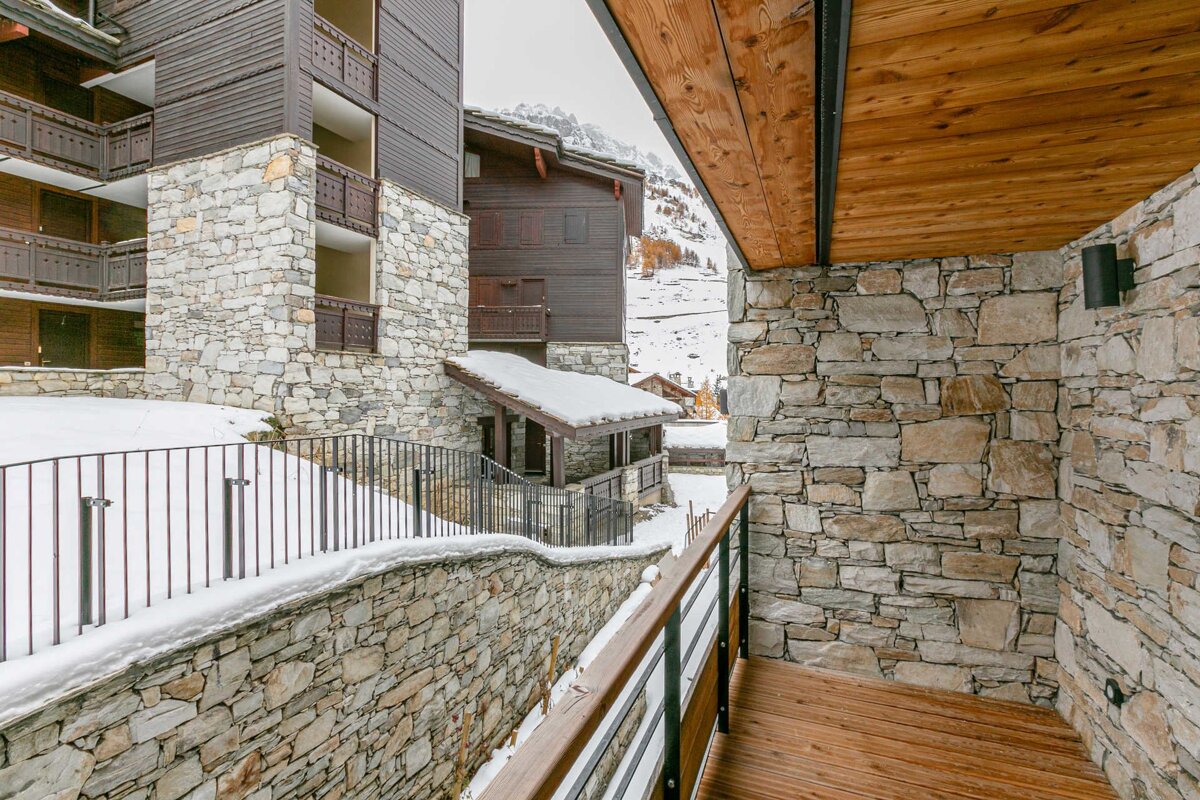 A snowy mountain resort with multiple stone and wood chalets, including a balcony with a wooden deck and railings. Distant snowy peaks visible.