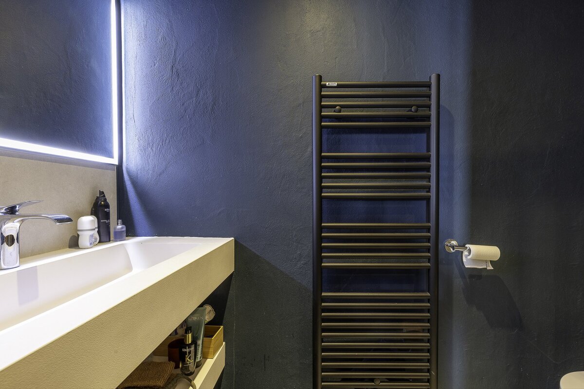 A modern bathroom features dark blue walls, a white trough sink with a backlit mirror, and a black heated towel rack.