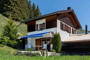 A two-story alpine chalet with a white lower level, wooden upper level, and a blue awning, nestled on a grassy hill with pine trees.