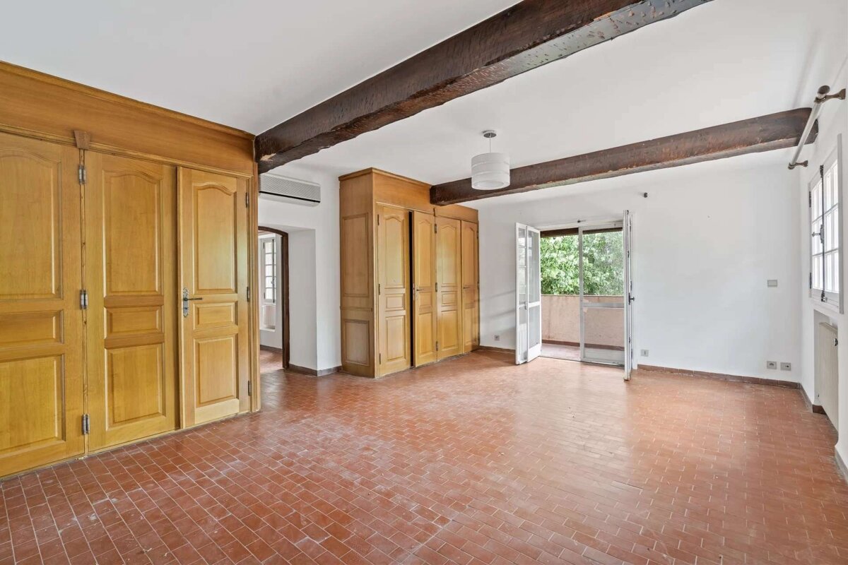 Empty room with terracotta tiled floor, exposed wooden beams, and light wood built-in wardrobes. An open balcony door reveals lush greenery.