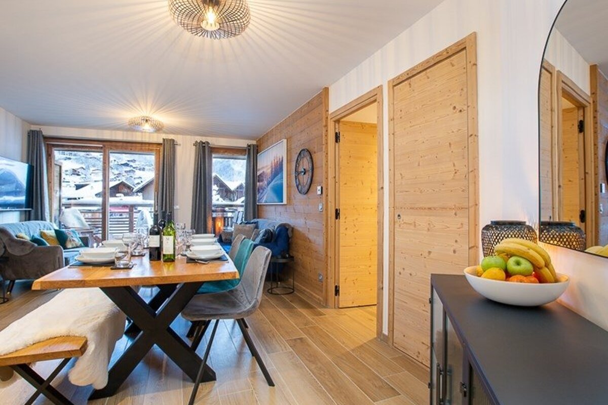 Cozy wooden chalet interior with a set dining table, modern decor, a fruit bowl, and snowy mountain views outside.