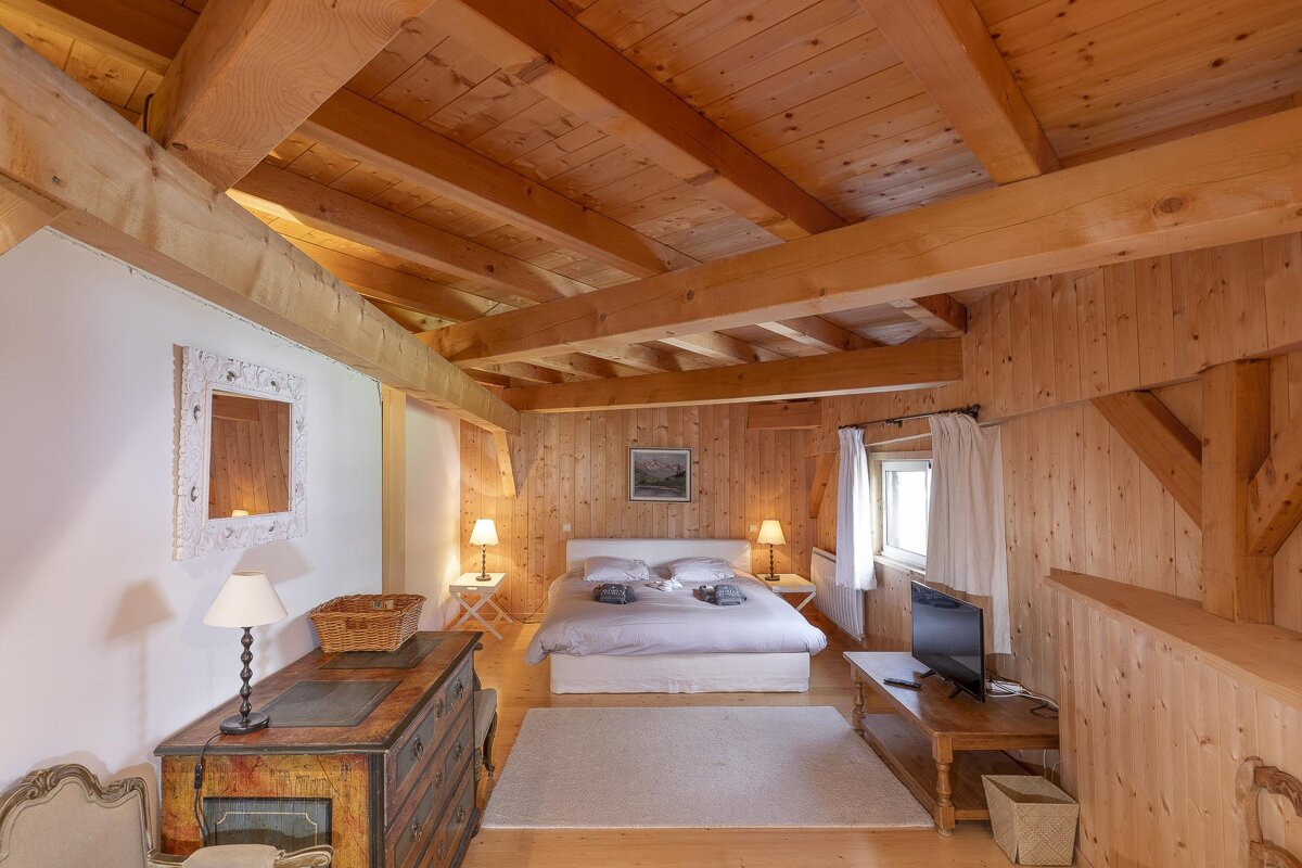 A rustic bedroom with extensive wood paneling, exposed beams, a comfortable bed, lamps, a dresser, and a TV, creating a warm ambiance.