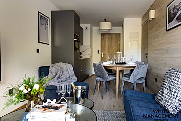 Modern open-plan living area with blue sofas, velvet dining chairs, a dark kitchenette, and wooden accents. Cozy decor includes flowers, champagne, and mountain art.