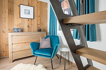A cozy room featuring a teal velvet armchair, wooden paneled walls, and a modern wood-and-metal staircase in the foreground.