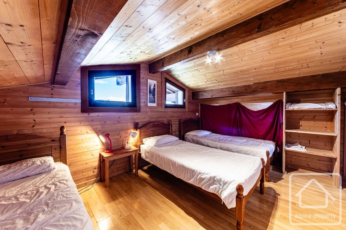 A rustic, wood-paneled attic bedroom with multiple single beds, a slanted ceiling, bright windows, built-in shelves, and a draped red fabric.