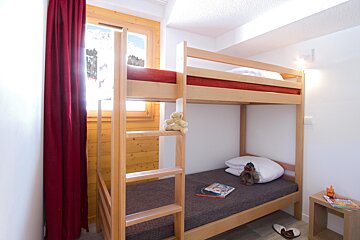A bright room with wooden bunk beds and red accents. A window overlooks snowy mountains, while toys and books suggest a child's space.