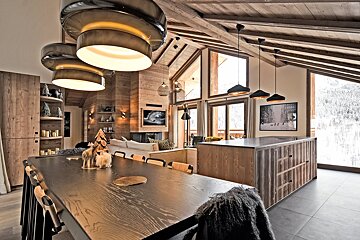 Modern alpine chalet interior featuring a wooden ceiling, chic pendant lights, dining table, kitchen island, and a snowy winter view.