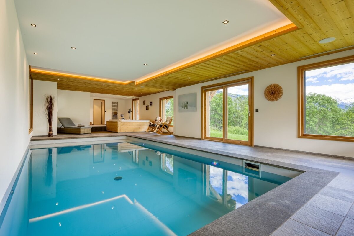 Bright indoor swimming pool with a wooden ceiling, large windows showing a lush mountain view, and a nearby lounge area.