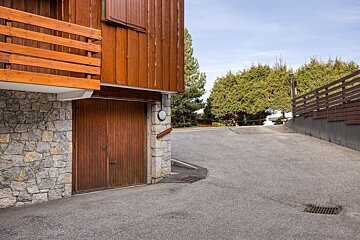 A rustic building with a stone base, wooden facade, and garage sits next to an uphill asphalt road. Green trees line the road under a blue sky.