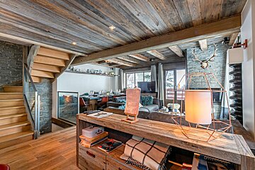 Rustic chalet interior with wooden ceilings, stone walls, staircase, living and dining areas, fireplace, and large windows.