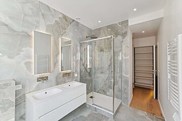 Modern bathroom featuring elegant marble walls, a double vanity with backlit mirrors, a glass shower, and built-in shelves.