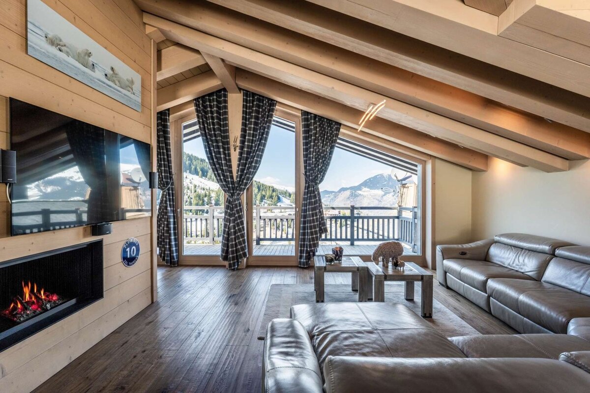 Cozy chalet living room featuring a TV, fireplace, leather sofas, wooden beams, and a panoramic balcony view of snowy mountains.