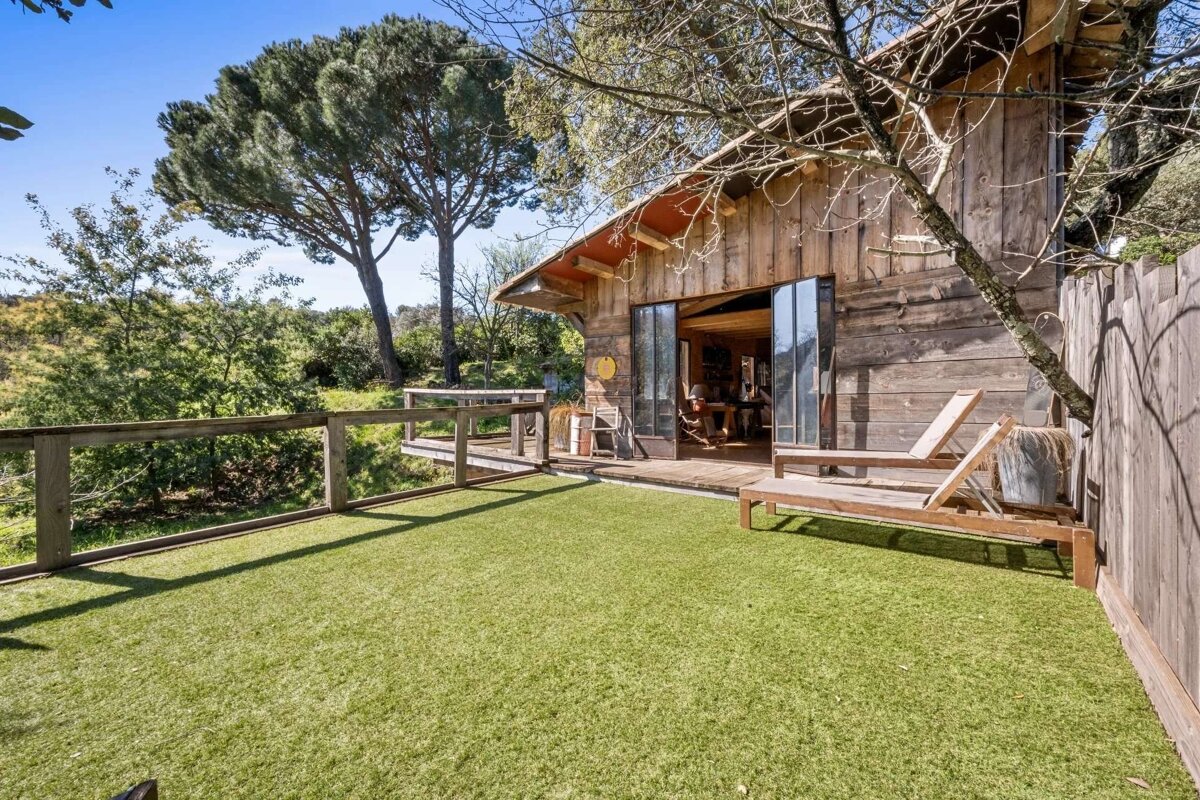 A rustic wooden cabin with a large artificial grass deck, two lounge chairs, and a natural tree-filled background under a clear blue sky.