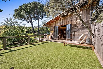 A rustic wooden cabin opens to an artificial grass patio with two lounge chairs, overlooking a green landscape under a sunny blue sky.