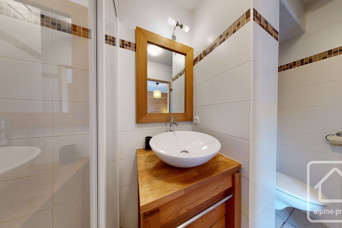 Modern bathroom with white tiled walls, brown mosaic accent, wooden vanity with a vessel sink, mirror, shower, and toilet.