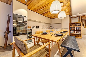 A stylish modern kitchen and dining area featuring a warm wooden ceiling, white cabinetry, a set dining table, built-in appliances, and vintage skis.