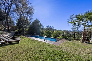 A sunny outdoor swimming pool surrounded by a green lawn, lounge chairs, lush trees, and distant hills under a clear blue sky.