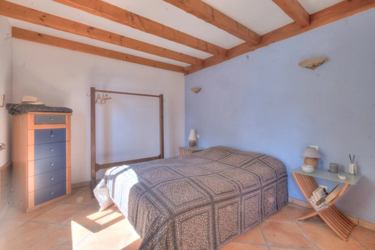 A bedroom with a blue accent wall, exposed wooden ceiling beams, terracotta tiles, a patterned bed, a dresser, and a clothing rack.