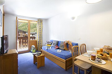 A bright studio apartment featuring a blue sofa, dining area with breakfast, and a balcony overlooking a mountainous village, all on a blue carpet.