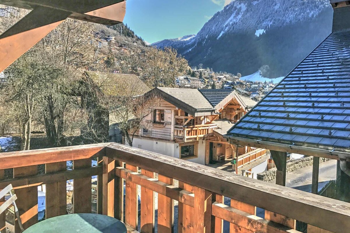 View of an alpine village with rustic wooden chalets and snowy mountains, taken from a wooden balcony.