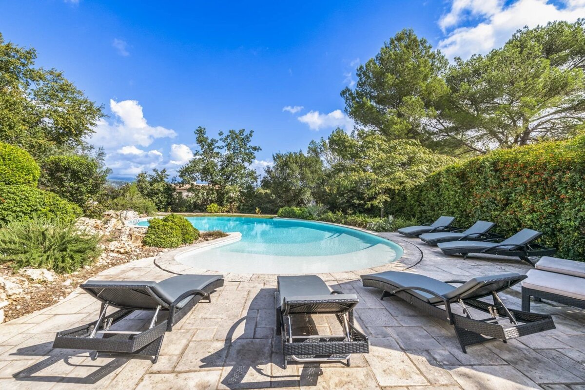 A curvy blue swimming pool with grey lounge chairs on a stone patio, surrounded by lush green trees and bushes under a bright blue sky with clouds.