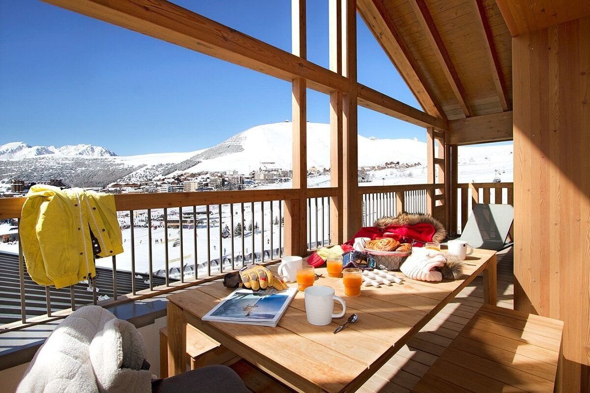 A cozy wooden balcony breakfast with orange juice and pastries, overlooking a stunning, sunny ski resort and snow-covered mountains.
