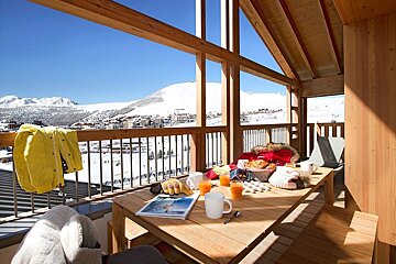 A cozy wooden balcony breakfast with orange juice and pastries, overlooking a stunning, sunny ski resort and snow-covered mountains.
