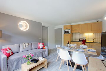 A modern open-plan living area with a comfy gray sofa, dining table set for dinner, and a well-lit kitchenette, all in a bright, neat space.