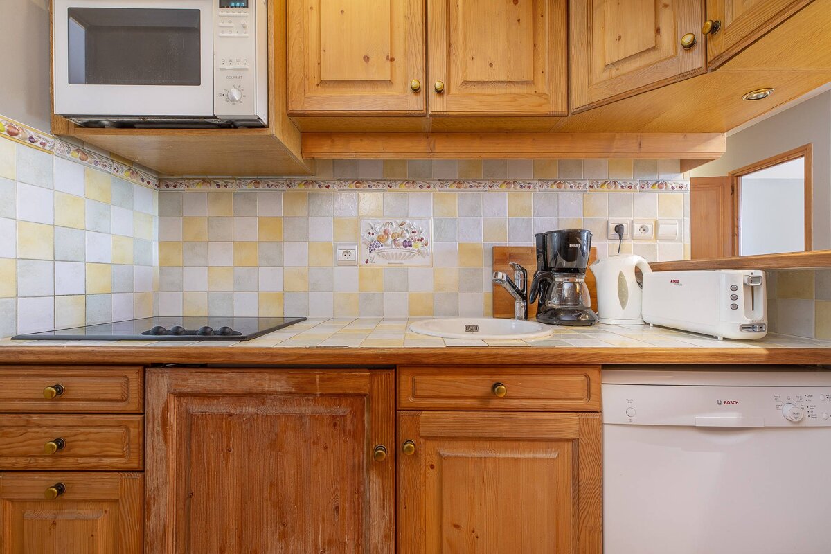 A compact kitchen with light wood cabinets, checkered yellow & white tile backsplash, microwave, stovetop, sink, coffee maker, kettle, toaster, and dishwasher.