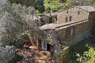 A rustic stone house complex with terracotta roofs and a sunny, tree-shaded patio, nestled among lush green hills.
