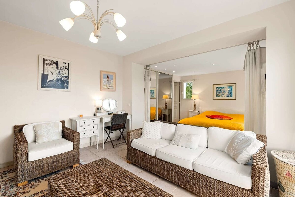 A bright, open-plan living room with wicker furniture, white cushions, and a dressing table, leading into a bedroom with a vibrant yellow bed.