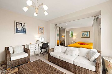 A bright, open-plan living room with wicker furniture, white cushions, and a dressing table, leading into a bedroom with a vibrant yellow bed.