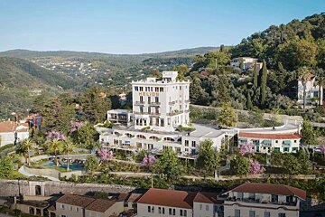 The elegant white Helios hotel is nestled on a lush green hillside with blooming trees, a pool, and distant mountains under a clear blue sky.